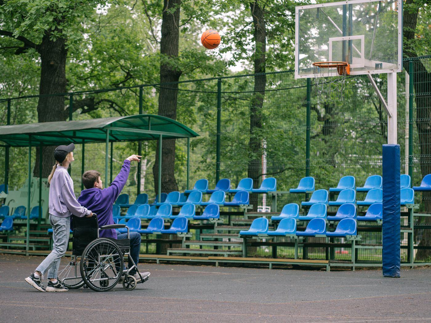 A support worker cheering on a young man in a wheelchair as he shoots a basketball at an outdoor court